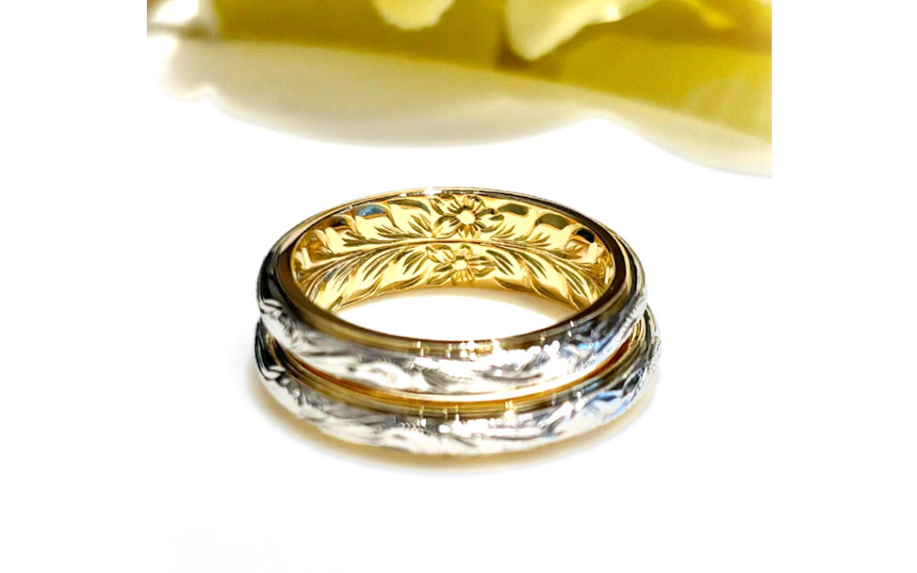 お選び頂きましたリングは、トップとベースに2色の異なる素材を組み合わせた『ツートーンリング』です。
トップには白く美しく輝く「プラチナ」を、ベースには華やかに輝く「18金イエローゴールド」のコンビネーションをお選び頂きました。
さらに、ハワイ伝統の手彫りを、リングの表面と内側の両面ともお楽しみ頂ける、特別な『両面彫り』にてお作りいただきました。
&nbsp;
表面には、波のデザインの〝永遠に受け継がれる想い〟という意味のある「ヘリテイジ」。
また、内側には、2つのリングを重ね合わせると神々が宿るとされる神聖な葉「マイレ」のデザインが完成する〝絆〟の意味を持つ「ピリナ」が、ハワイの熟練の彫師の手によって刻まれました。
マイレの葉は、ハワイでは古来から重要な儀式で用いられており、大切な人との絆を象徴するとされています。
