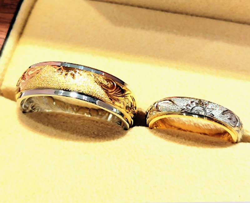 リングの表面は手彫りとシンプルさを両方楽しめる、半周切替えの2Wayデザインに。
&nbsp;
N様は〈生命力・成功〉を表す「太陽」を、K様は〈一途な愛、情熱〉の意味を持つ「向日葵」を半分ずつ彫りこみ、その両側に絶え間なく幸せを運び込む〈永遠〉を象徴する波のデザイン「プリンセス」を。
&nbsp;
また、波の始まり部分に 釣り針のモチーフ「フィッシュフック」を波に見立て、おふたりのリングを重ね合わせるとハートになるように彫り込み、ペア感をしっかりと出されました。
「フィッシュフック」は〈幸運を掴んで放さない〉という意味を持っています。
&nbsp;
背景には砂模様の「サンド」加工をリング半周にお入れされ、メリハリをつけながら、彫りが立体的に浮かび上がるように仕上げられました。
